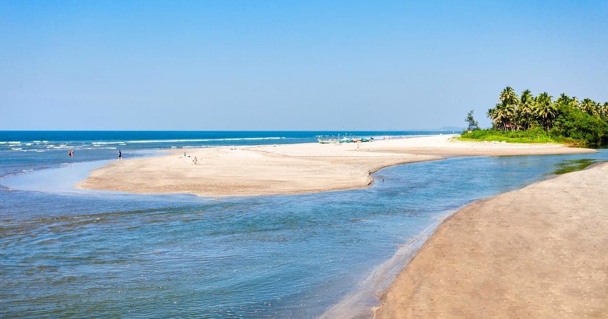 A wide sandy beach in Goa where a river meets the sea, with palm trees on the far shore